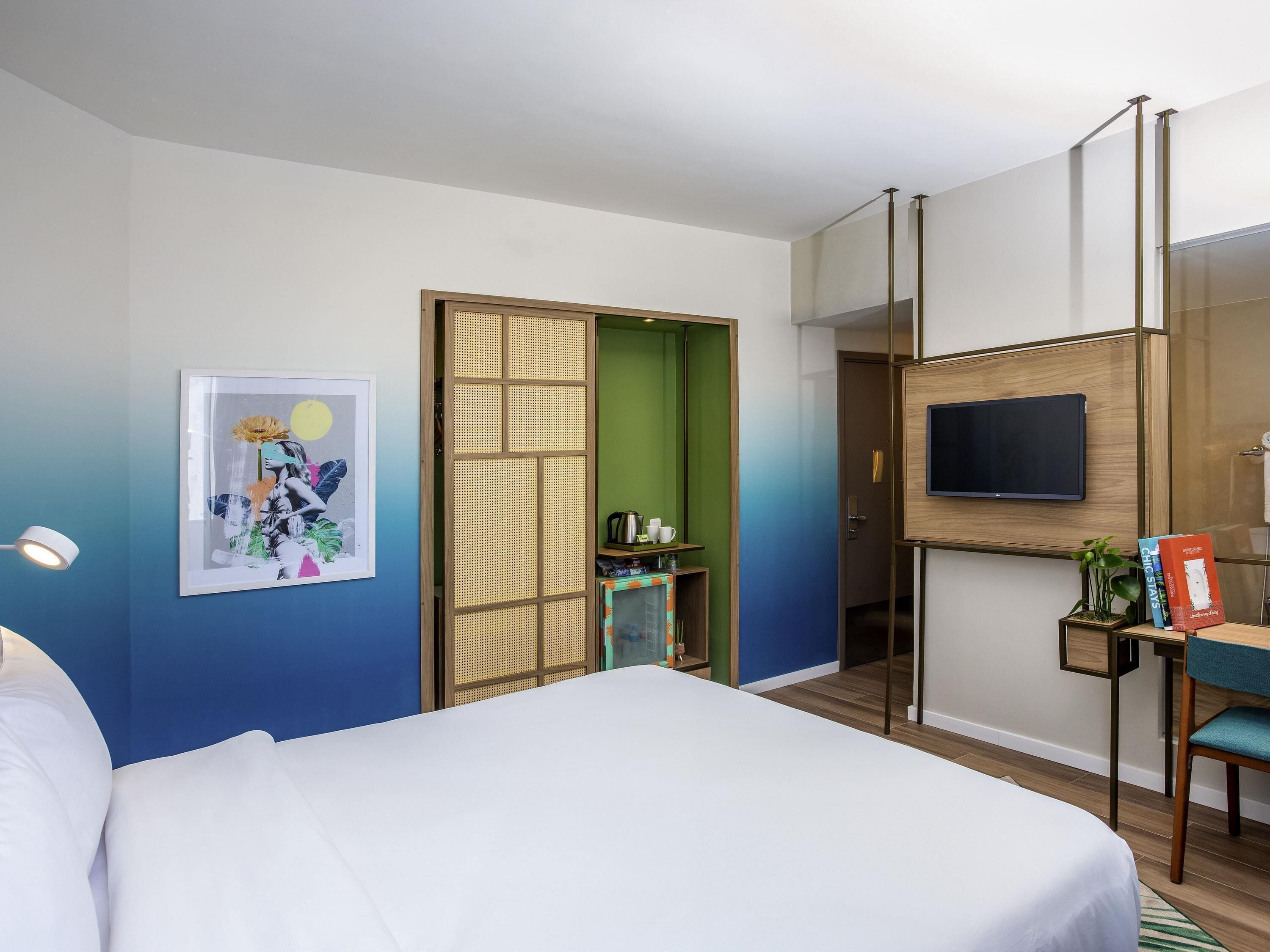 A hotel room with a bed, a TV mounted on the wall, a desk with a chair, and a decorative screen door.