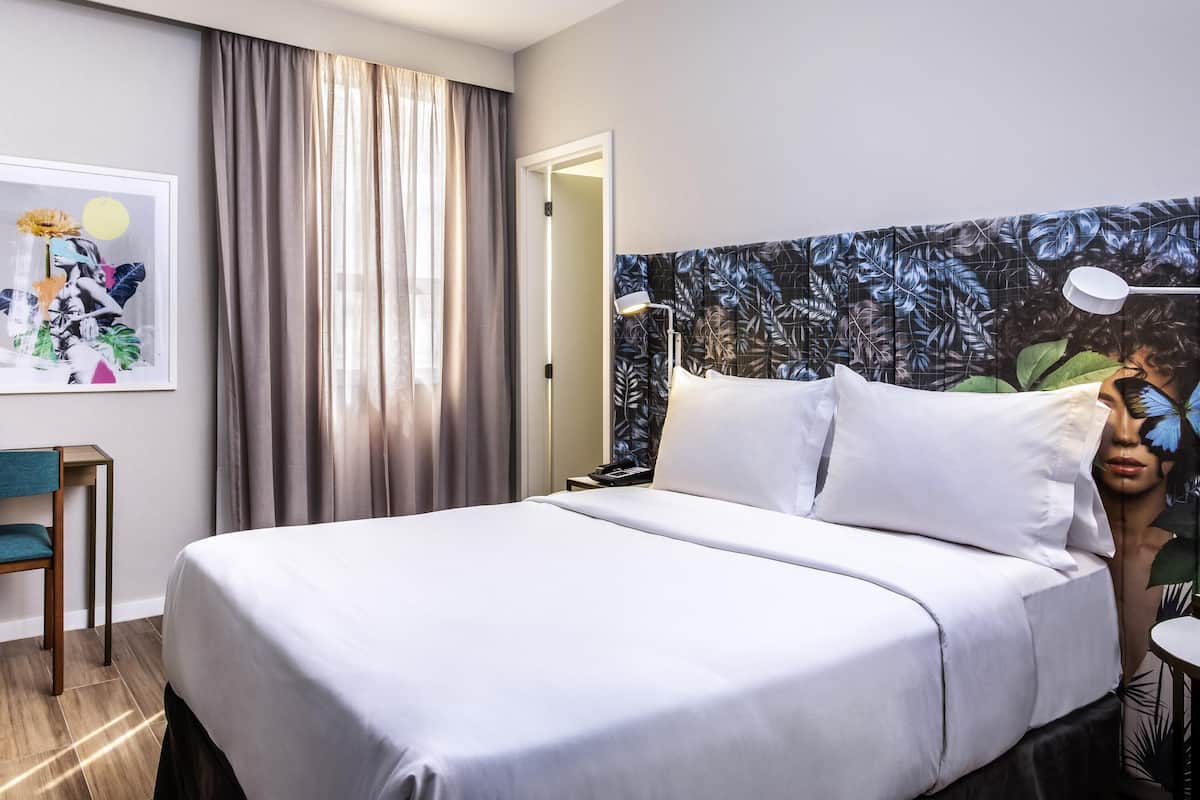 A hotel room with a bed, a desk, a chair, a nightstand, a lamp, and a large floral-patterned headboard.