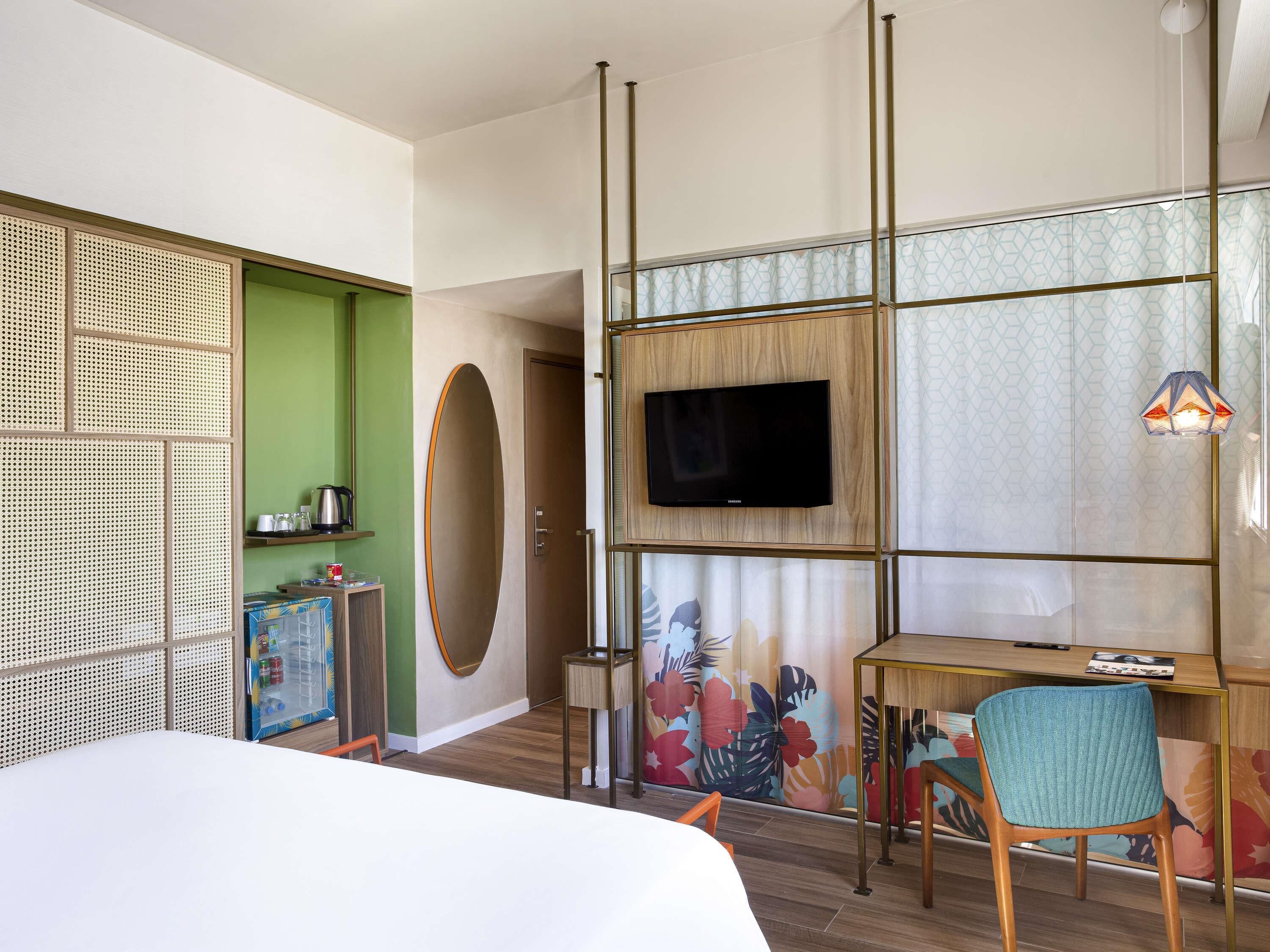 A hotel room with a bed, a TV, a desk with a chair, a decorative mirror, and a green accent wall.