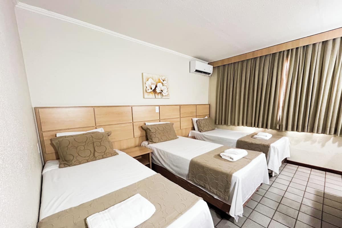 Habitación de hotel con tres camas individuales, un ventanal con cortinas, un aire acondicionado de pared y una obra de arte floral.