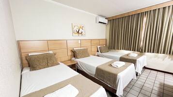 Quarto de hotel com três camas de solteiro, uma janela grande com cortinas, um ar condicionado fixado na parede e uma obra de arte floral.