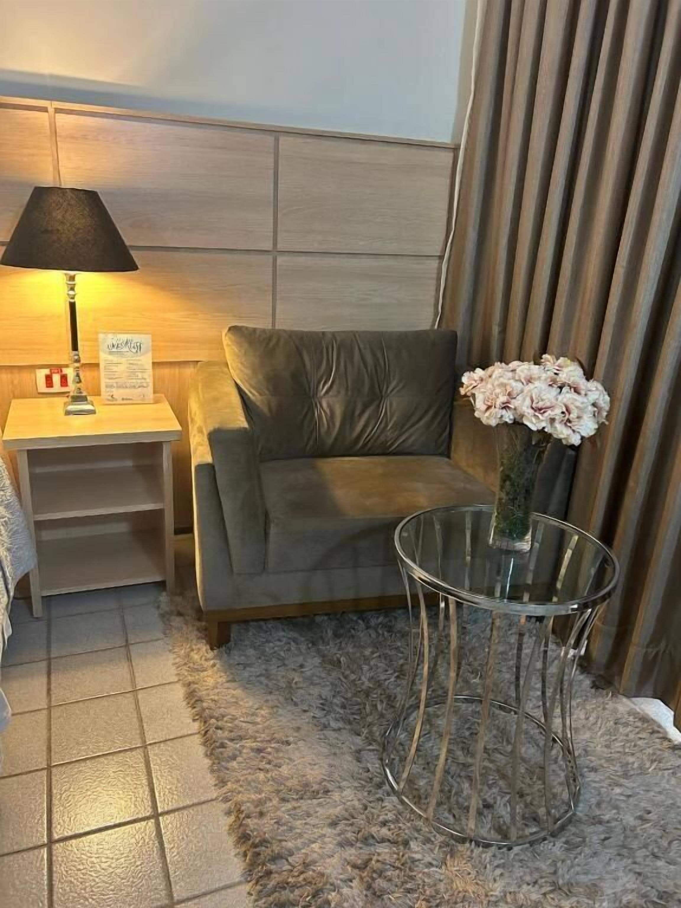 Una habitación con un sofá gris, una pequeña mesa de vidrio con un florero, una lámpara y un cartel en la mesita de noche.