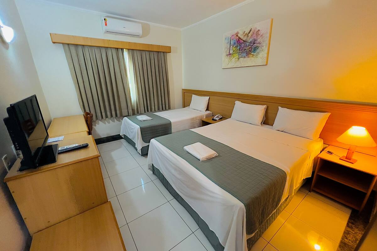 Habitación de hotel con dos camas, un escritorio con computadora, televisor, aire acondicionado, un cuadro en la pared y una lámpara de noche.