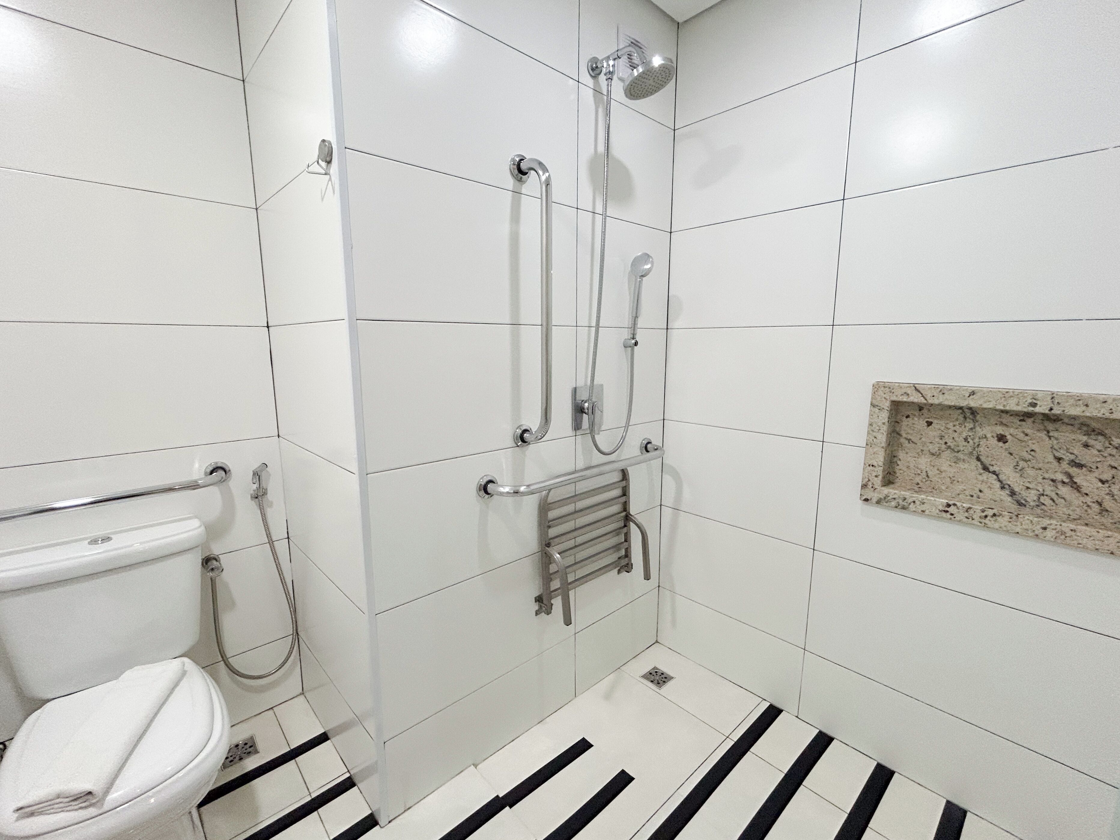 A bathroom with white tiles, a toilet, a shower with a handheld showerhead, and a towel rack.