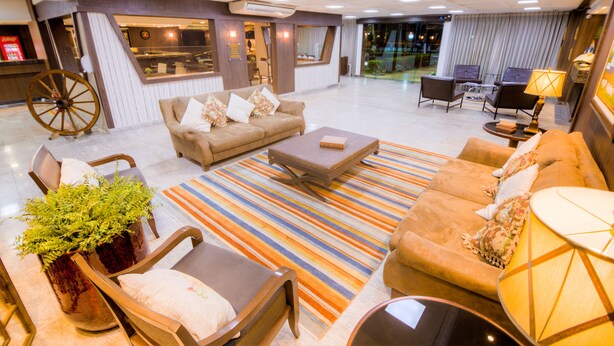 A spacious lobby with a large sofa set, a coffee table, and a striped rug. There is a large wooden wheel, a reception desk, and a seating area with chairs and a lamp. - Hotel Dan Inn Uberaba & Convenções by Nacional Inn (Uberaba)