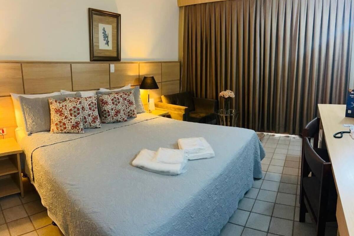 Una habitación de hotel con cama, almohadas, mesita de noche, lámpara, silla, escritorio y cortina.