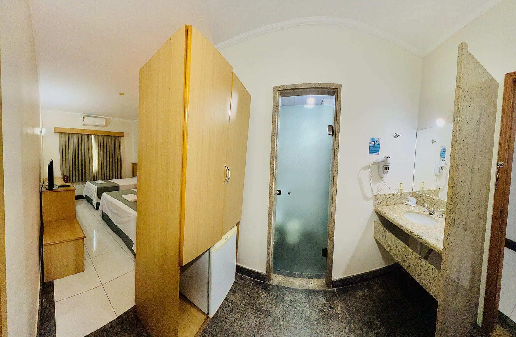 Ein Hotelzimmer mit Bett, einem Holzschrank, einem Badezimmer mit Waschbecken und Spiegel sowie einer Tür zu einem anderen Raum.