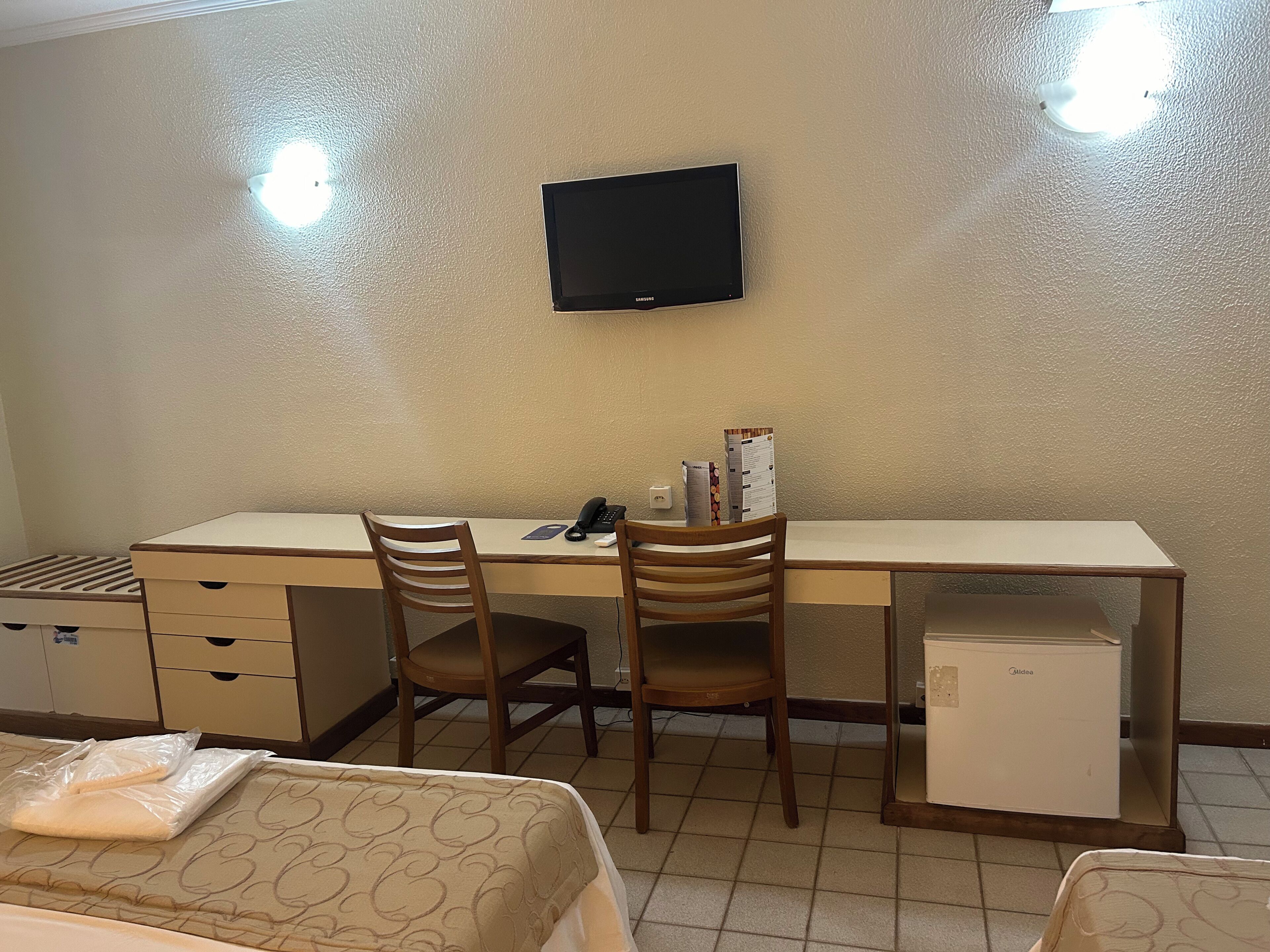 Habitación de hotel con escritorio, dos sillas, televisor, refrigerador pequeño y cama con colcha estampada.