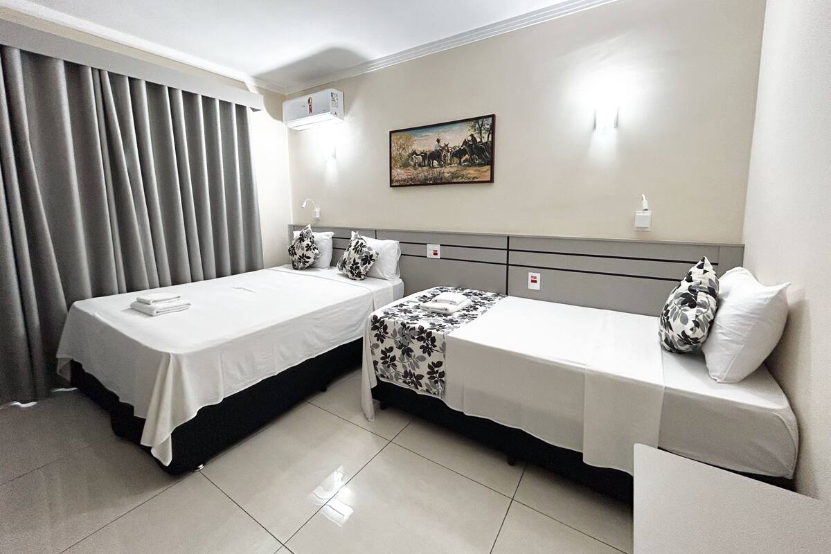 Habitación de hotel con dos camas individuales, un ventanal grande con cortinas, un cuadro en la pared y un aire acondicionado instalado en el techo.
