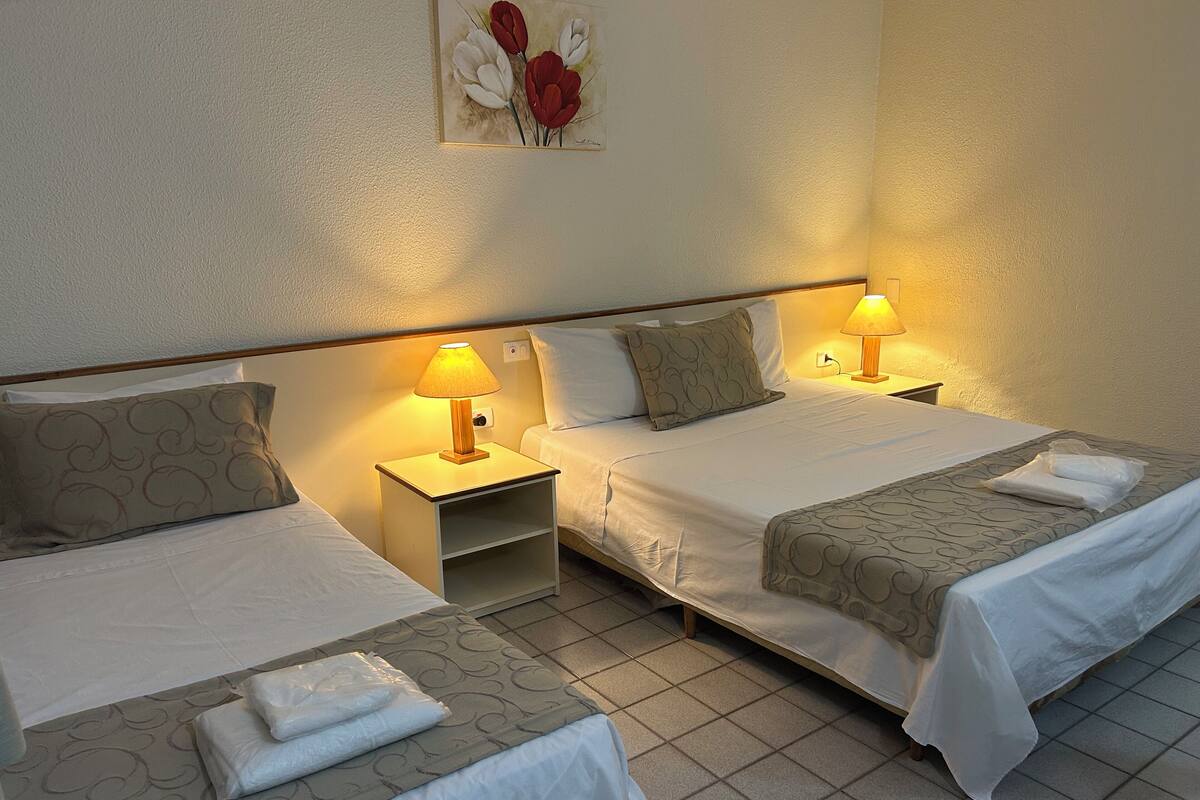 Habitación de hotel con dos camas, cada una con una almohada y una manta, una mesita de noche con una lámpara, y un cuadro en la pared.