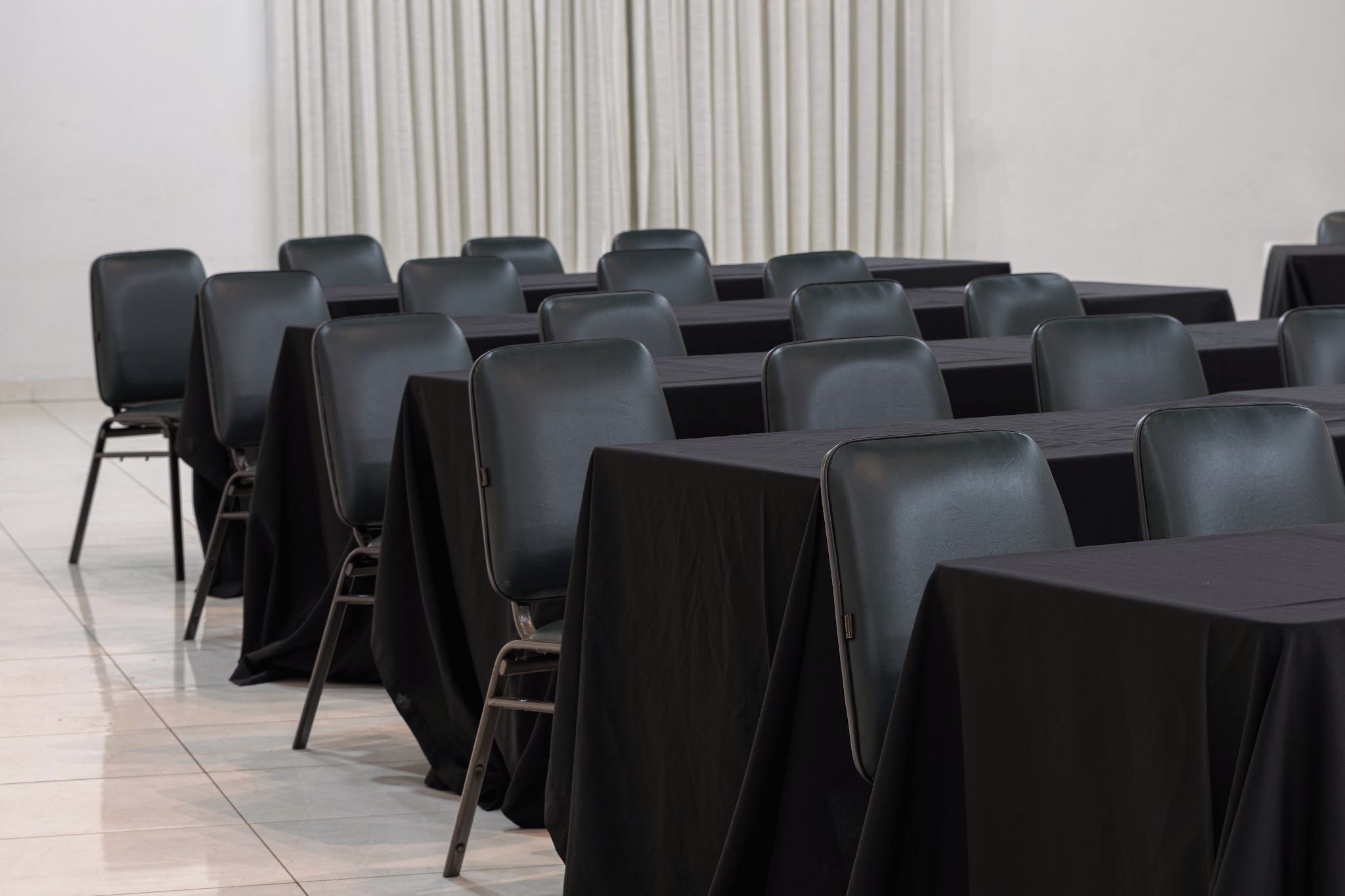 A conference room with rows of tables and chairs, a tiled floor, and a plain wall with vertical blinds.