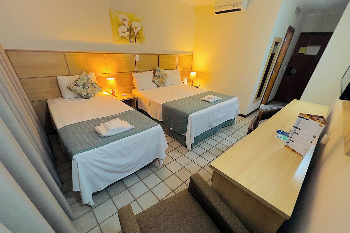 Habitación de hotel con dos camas, un escritorio y una silla. Hay un cuadro en la pared y un equipo de aire acondicionado sobre las camas.
