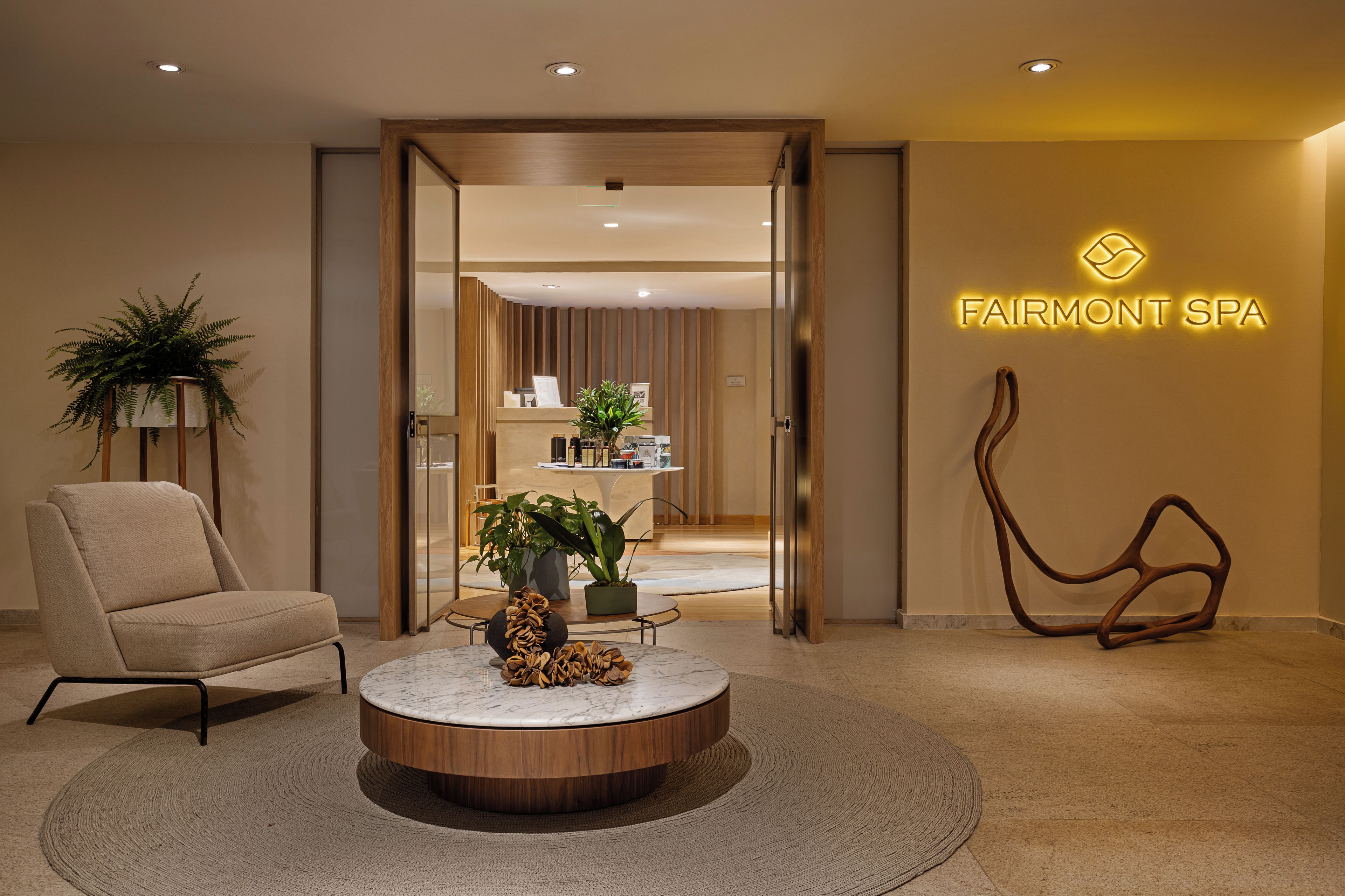 Zona de recepción con mostrador, una silla, una mesa redonda con una planta y un letrero en la pared para el Spa Fairmont.