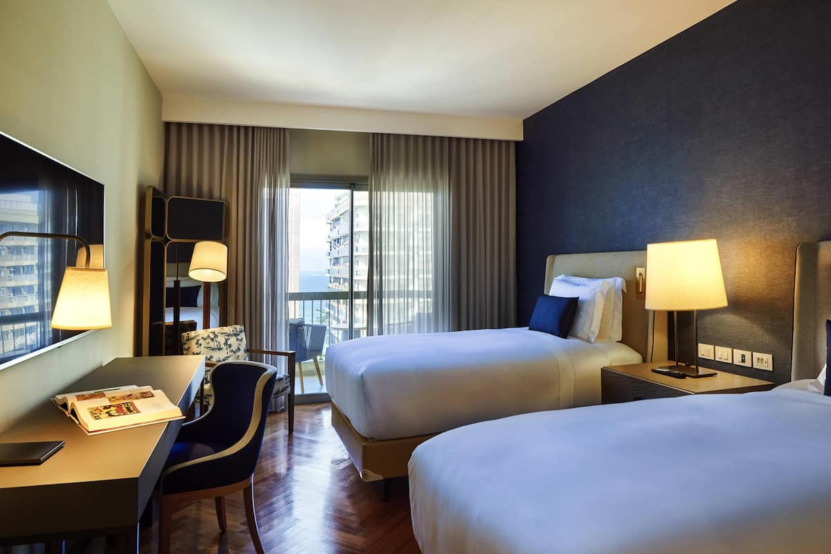 Ein Hotelzimmer mit zwei Betten, einem Schreibtisch mit Stuhl, einer Lampe und Blick auf ein Gebäude durch das Fenster.
