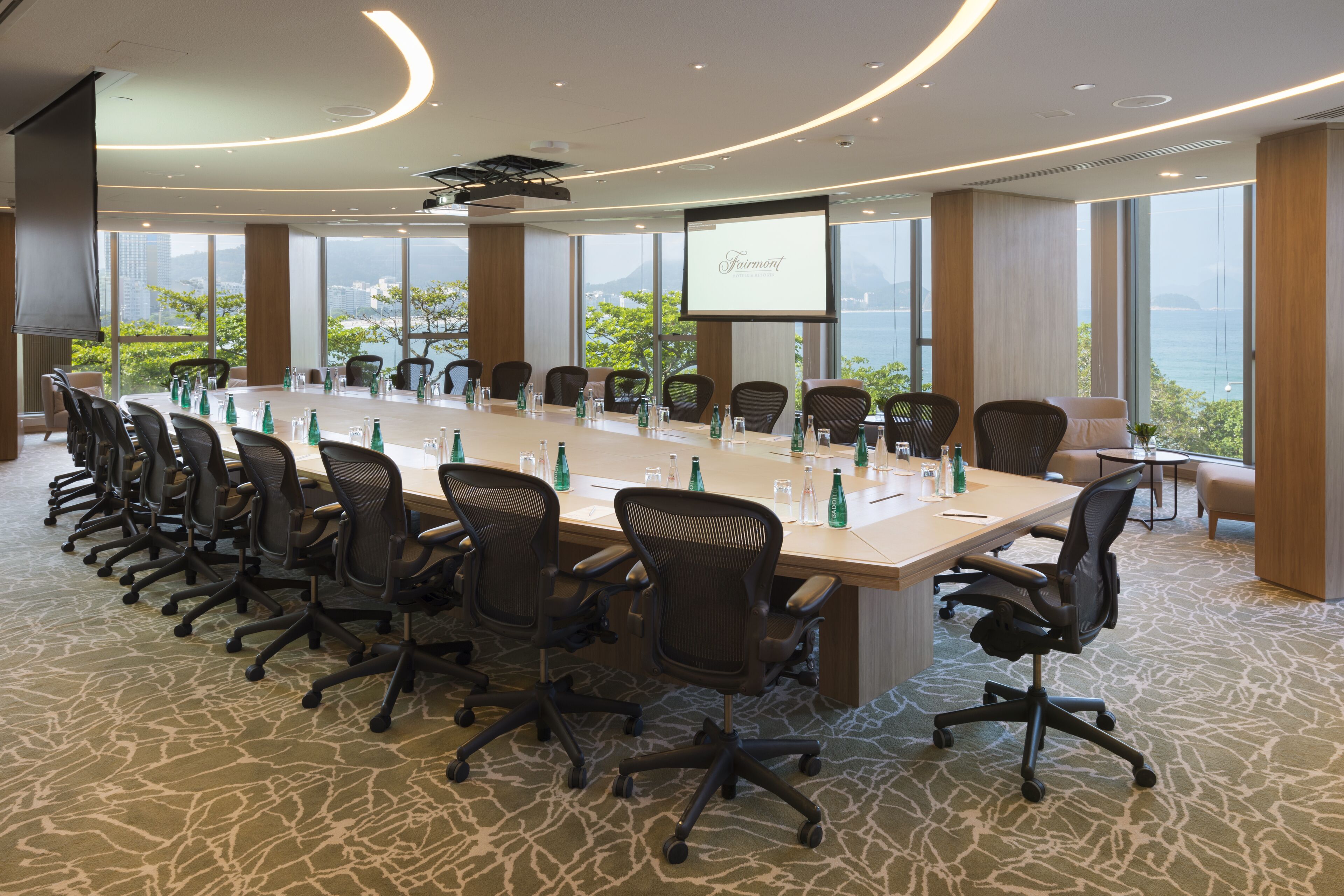 Une salle de conférence équipée d’une longue table, de chaises, d’un écran de projection et de grandes fenêtres offrant une vue dégagée.