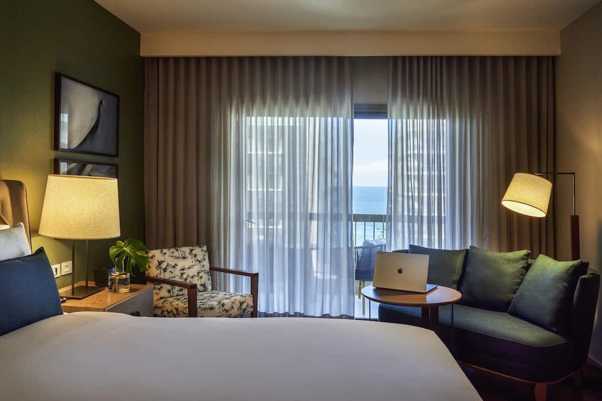 A hotel room with a bed, a desk with a laptop, a chair, a sofa, a lamp, and a view of the sea through large curtains.
