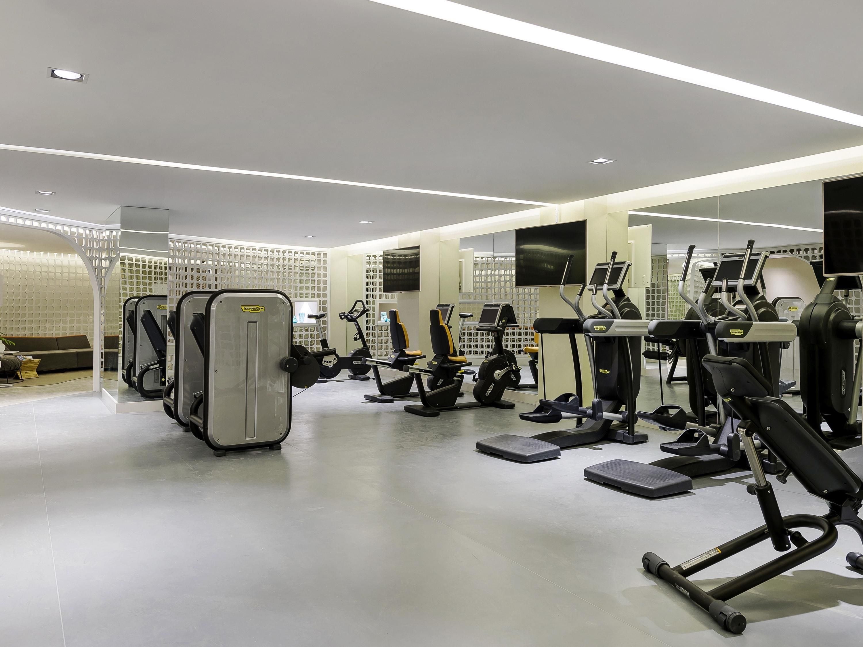Academia moderna com diversos equipamentos de exercício, um espelho grande e uma área de descanso com sofá.