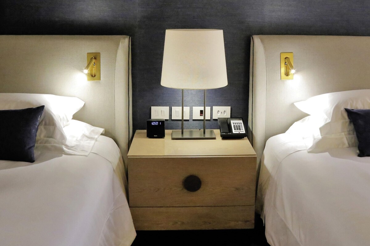 A hotel room with two beds, a bedside table with a lamp, a digital clock, and a telephone.
