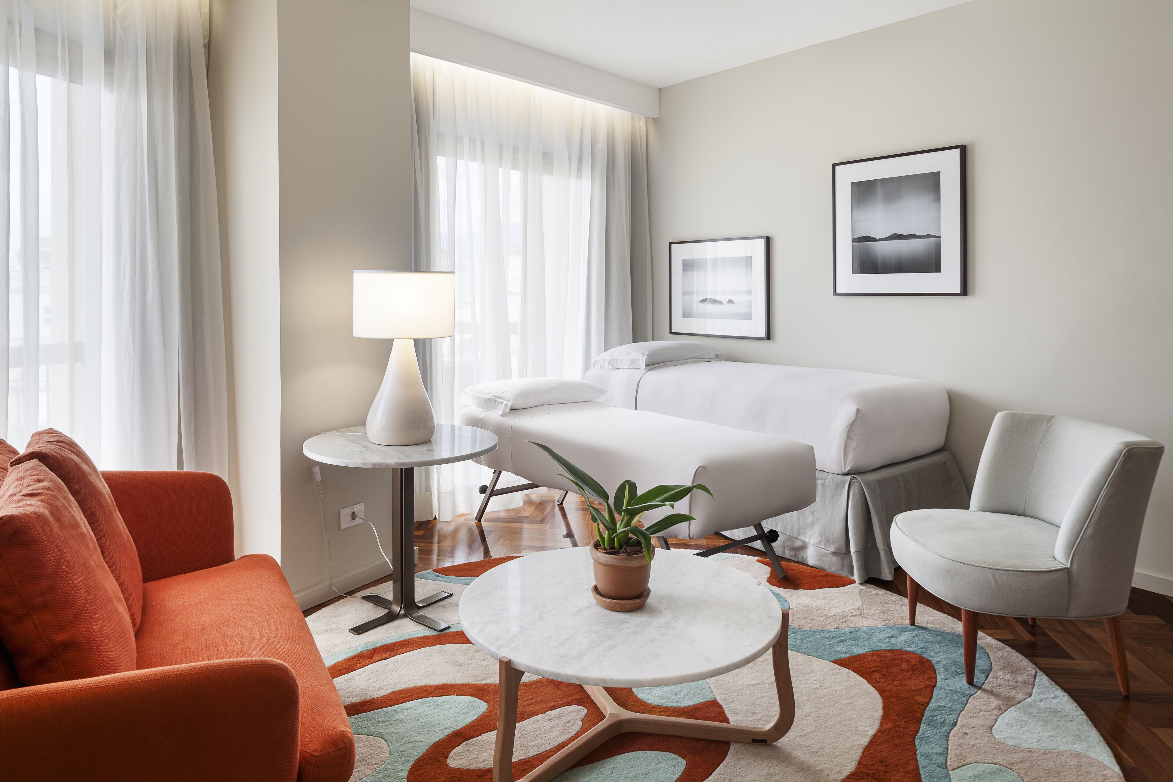 A hotel room with a bed, a sofa, a chair, a small table with a lamp, a potted plant, and two framed pictures on the wall.