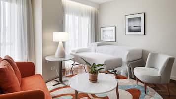 Habitación de hotel con cama, sofá, sillón, una mesita con lámpara, una planta en maceta y dos cuadros en la pared.