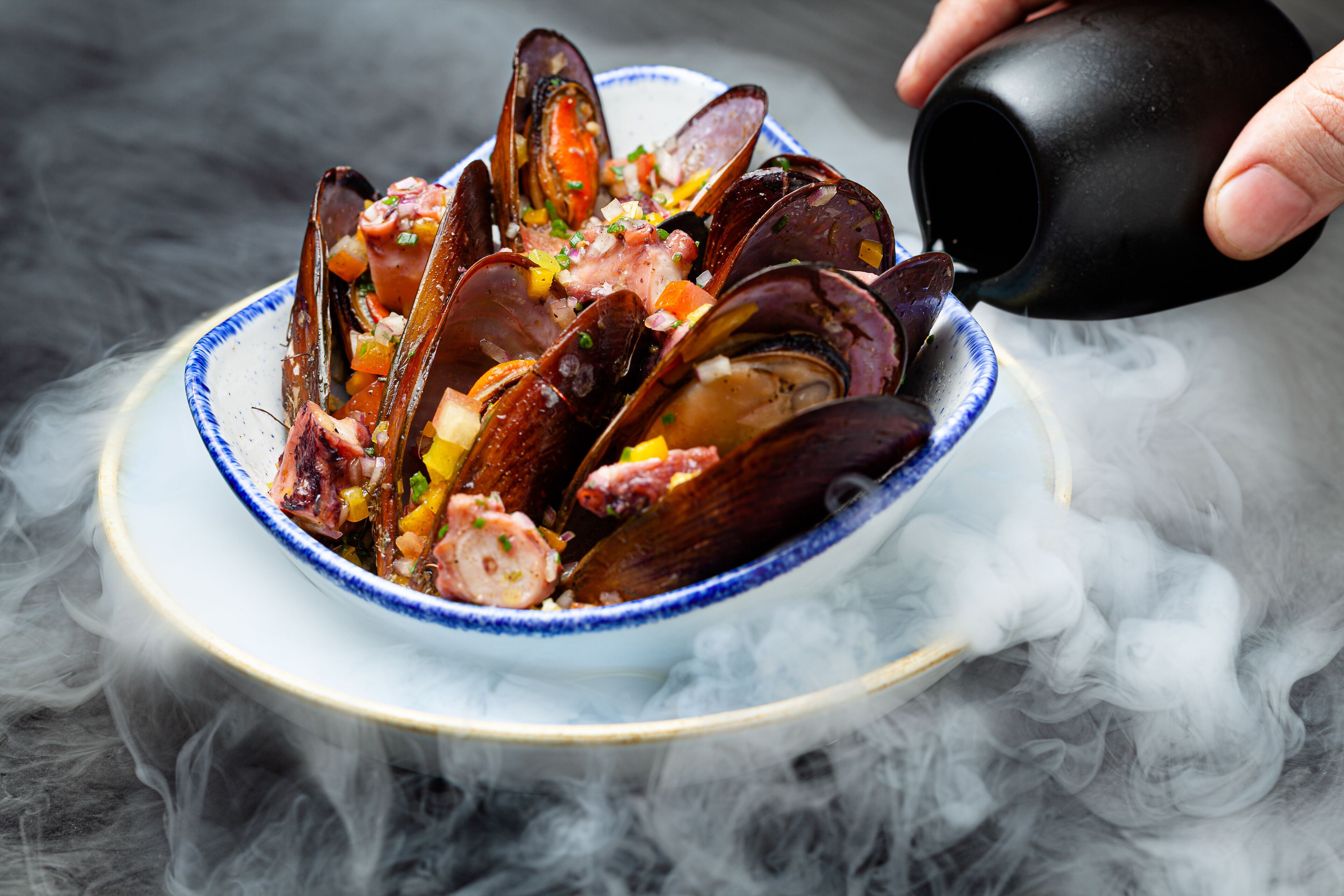 A bowl of mussels with a garnish, being poured from a black cup.