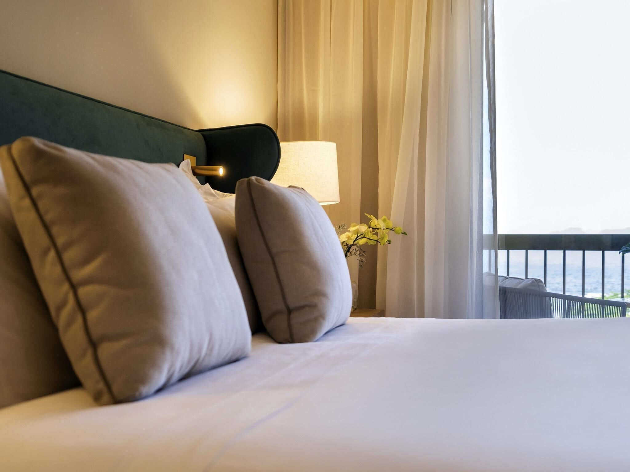 Un letto rifatto con cuscini, una lampada da comodino e vista sul mare attraverso le tende.