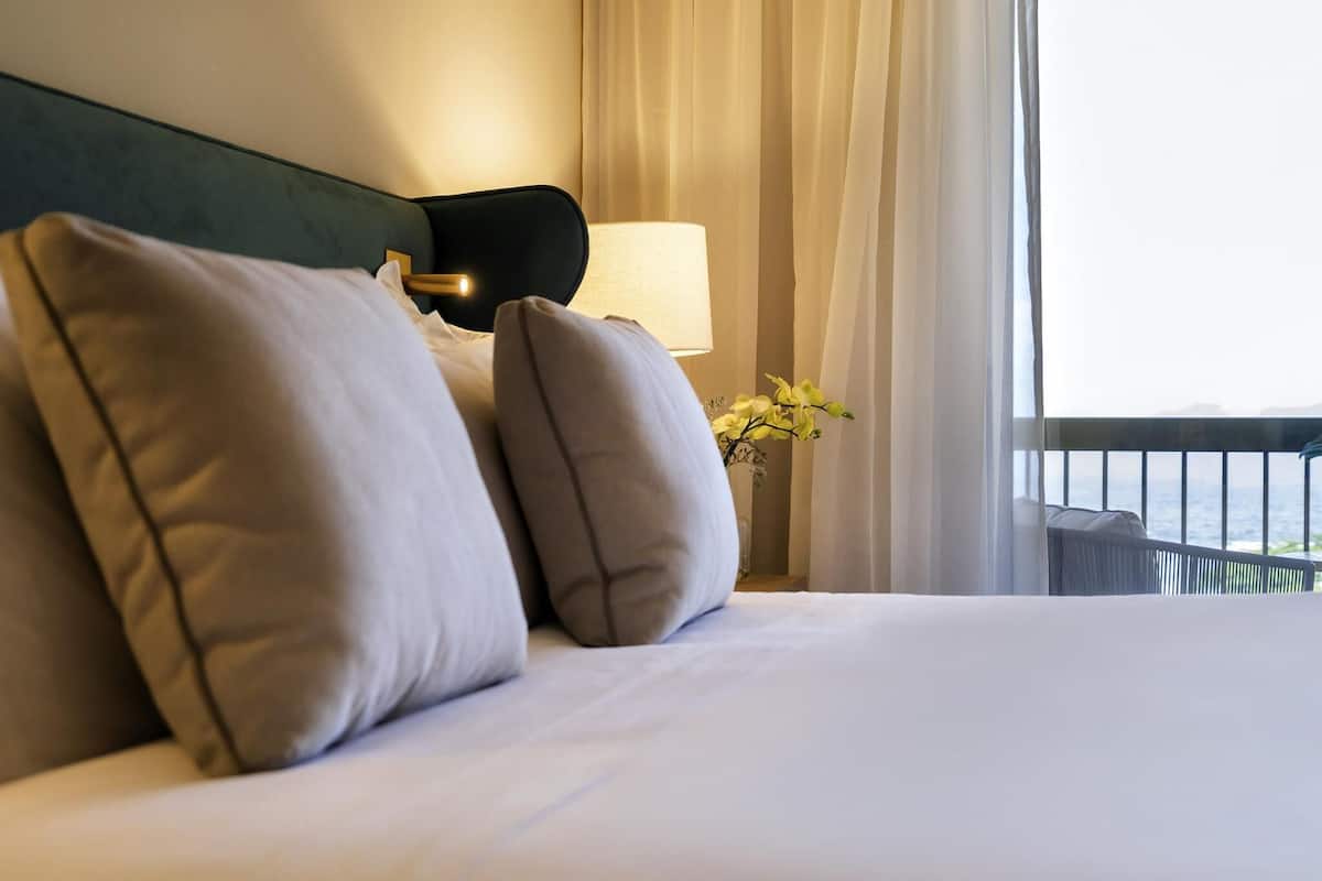 Un letto rifatto con cuscini, una lampada da comodino e vista sul mare attraverso le tende.