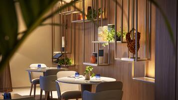 A modern dining area with round tables and chairs, wooden paneling, and shelves with potted plants and decorative items.