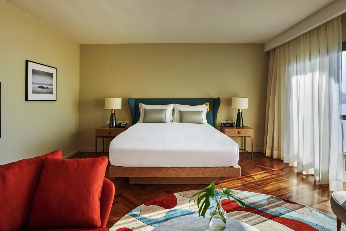 A hotel room with a large bed, two bedside tables with lamps, a red armchair, a patterned rug, and a framed picture on the wall.