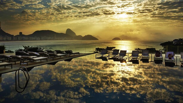 An infinity pool with lounge chairs facing a sunset over the ocean, with cityscape in the background. - Fairmont Rio de Janeiro Copacabana (Rio de Janeiro)