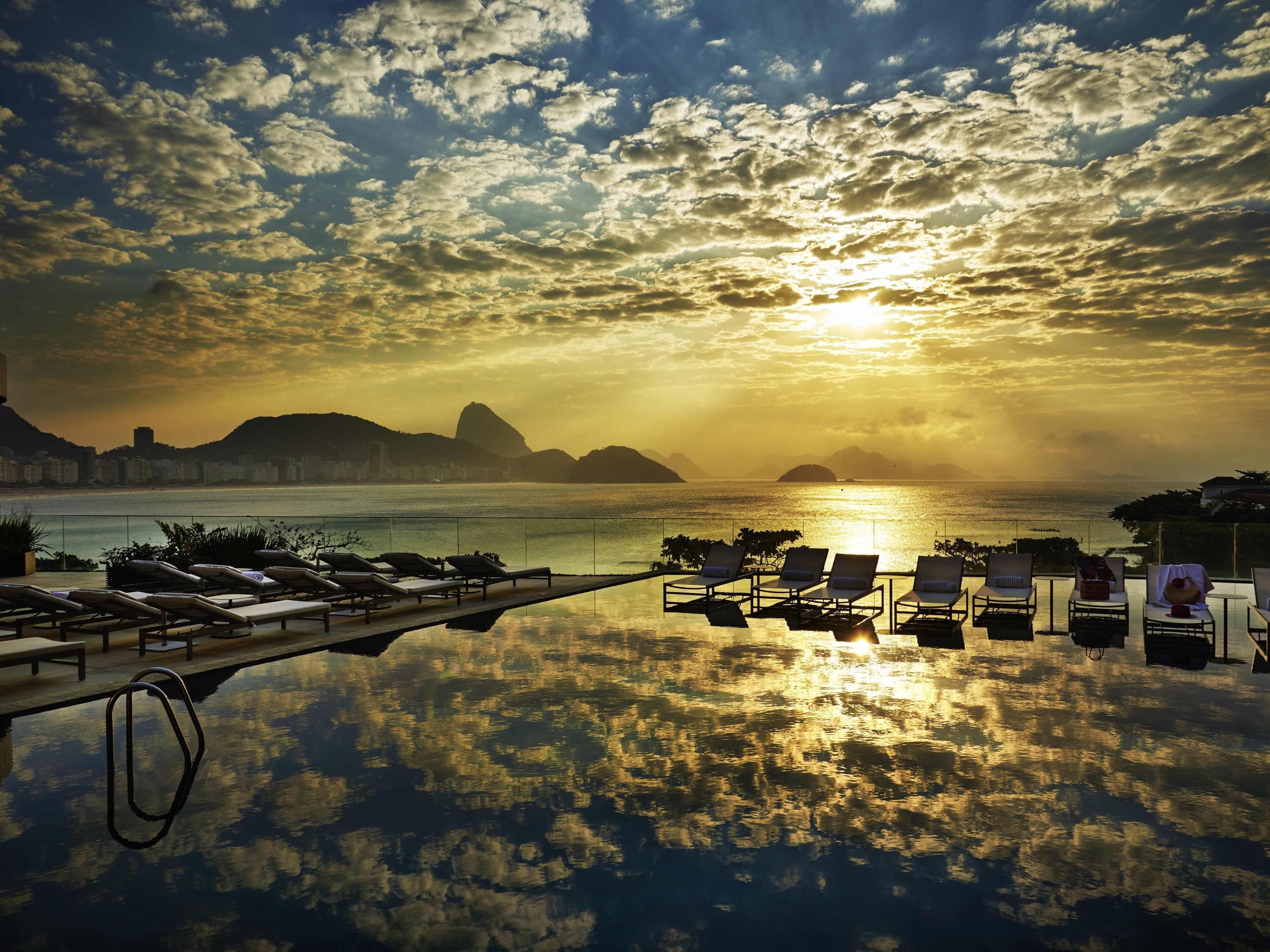 An infinity pool with lounge chairs facing a sunset over the ocean, with cityscape in the background.
