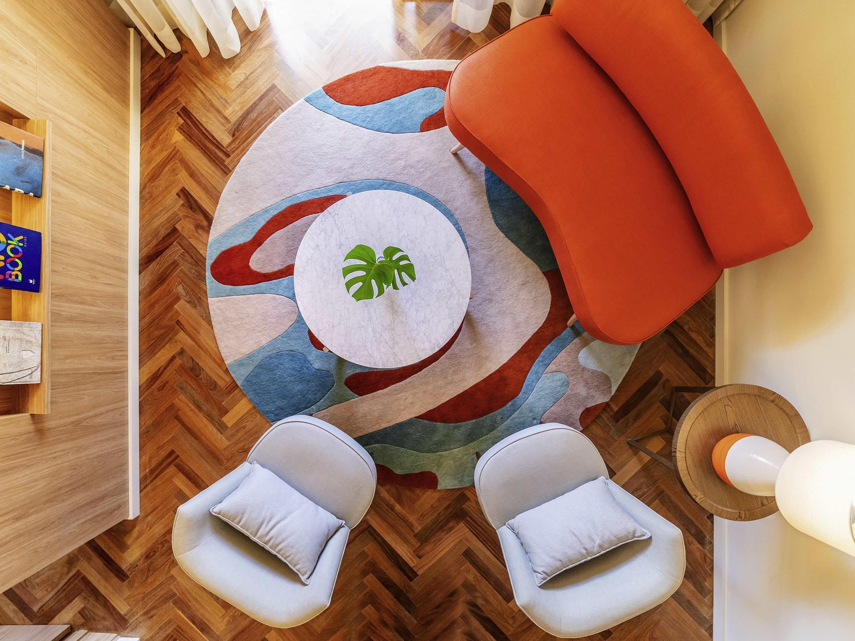 A room with a herringbone wooden floor, a round rug with a leaf pattern, two white armchairs, and a red ottoman.