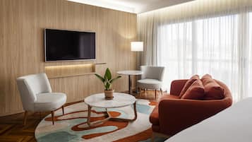 A modern hotel room with a flat-screen TV mounted on the wall, a round coffee table with a plant, two armchairs, a red sofa, and a large window with sheer curtains.