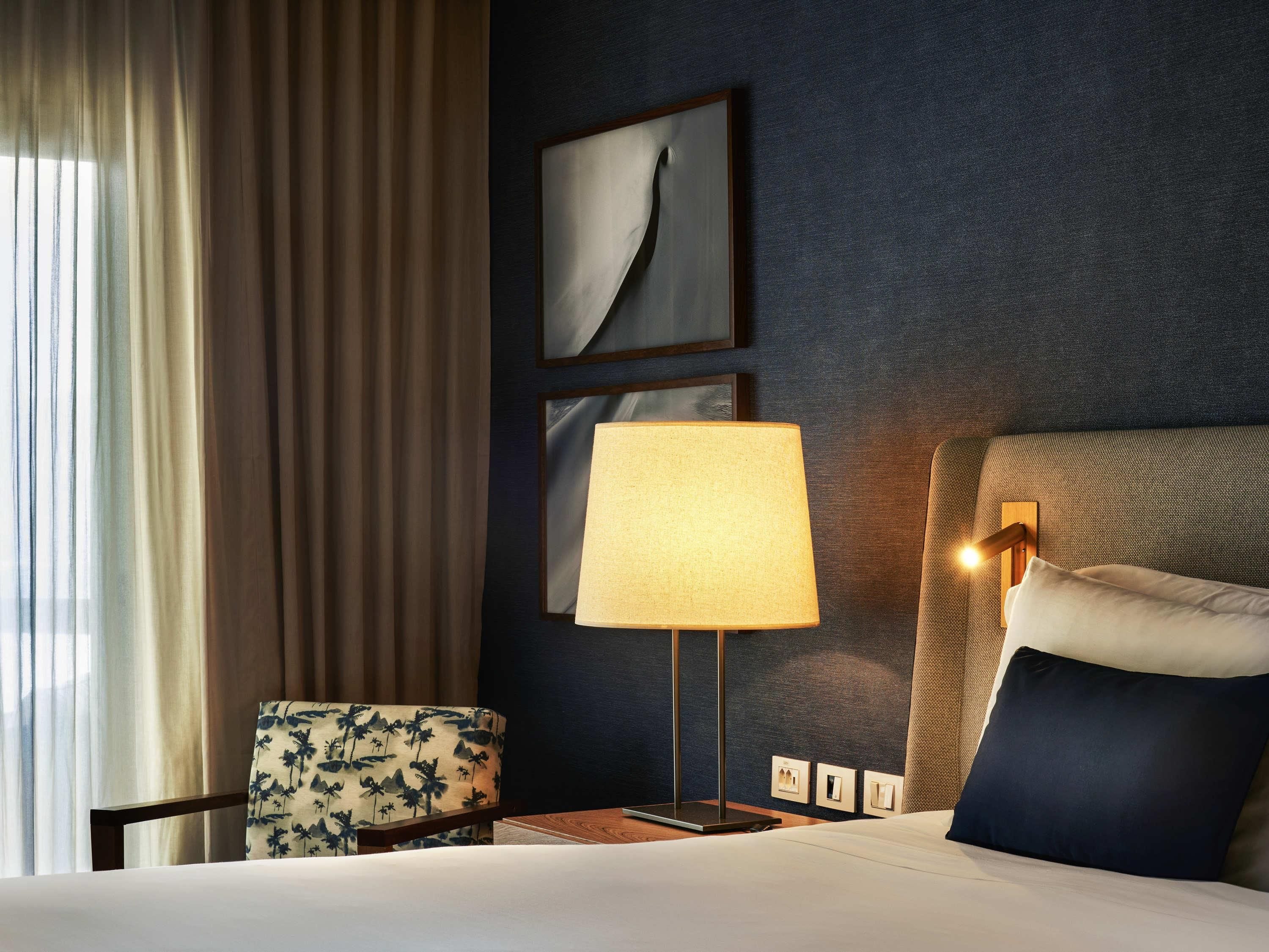 A hotel room with a bed, a desk, a chair, a lamp, and a wall-mounted light fixture.