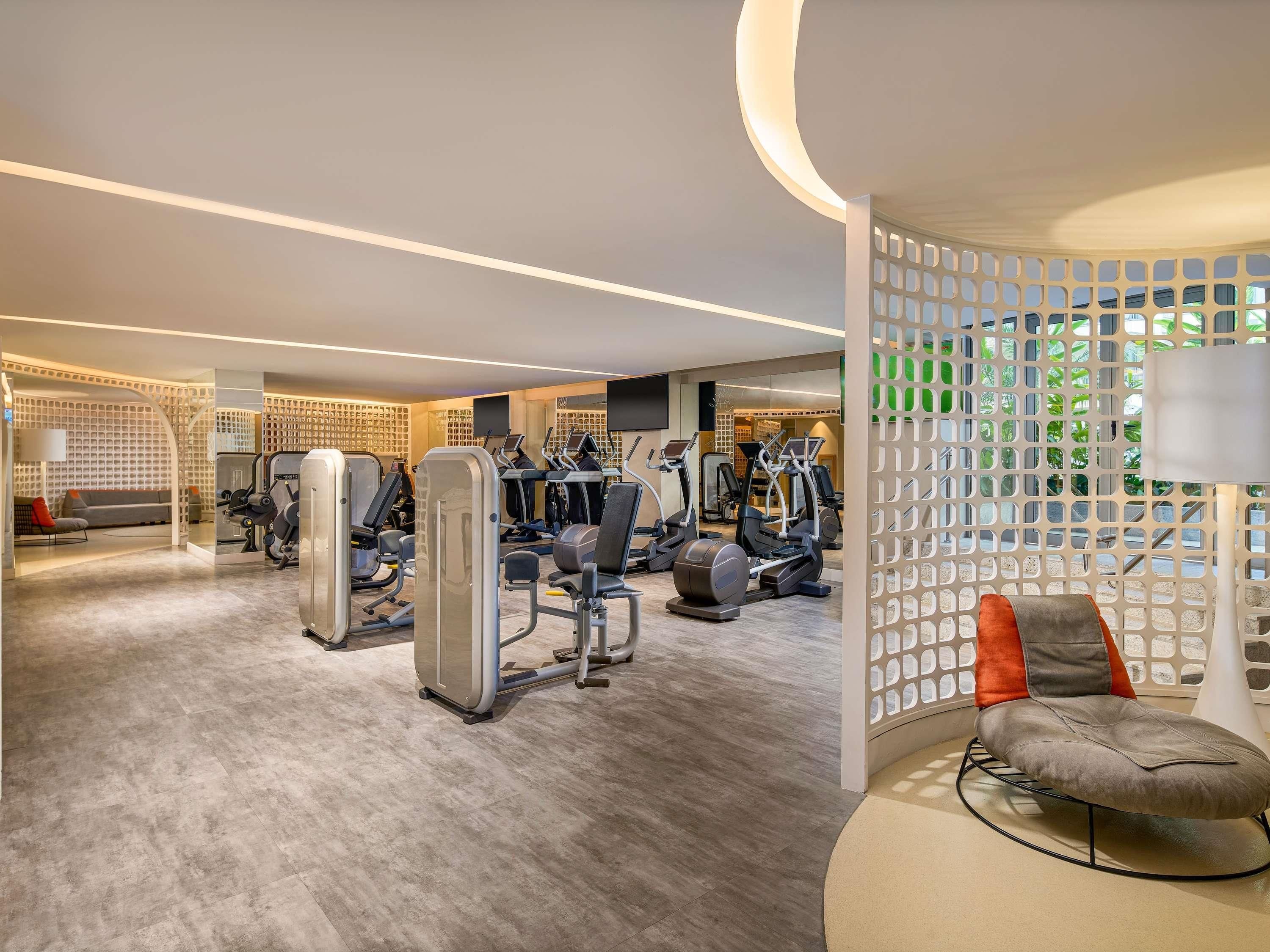 Uma academia moderna com diversos equipamentos de exercício, uma área de estar com sofá e cadeiras, e uma parede decorativa com padrão geométrico.