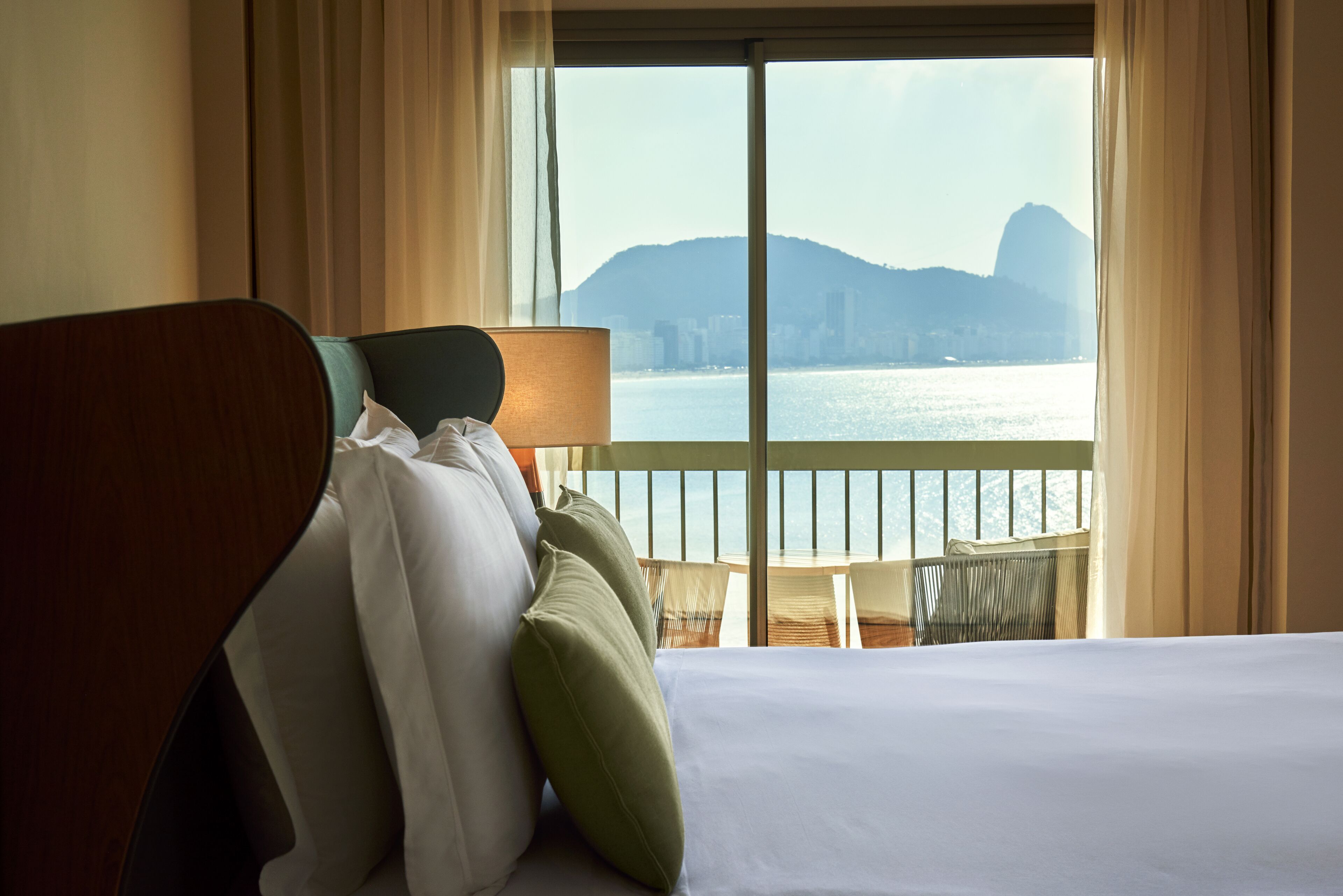 A hotel room with a bed, pillows, a nightstand, a lamp, and a view of a body of water and distant mountains.
