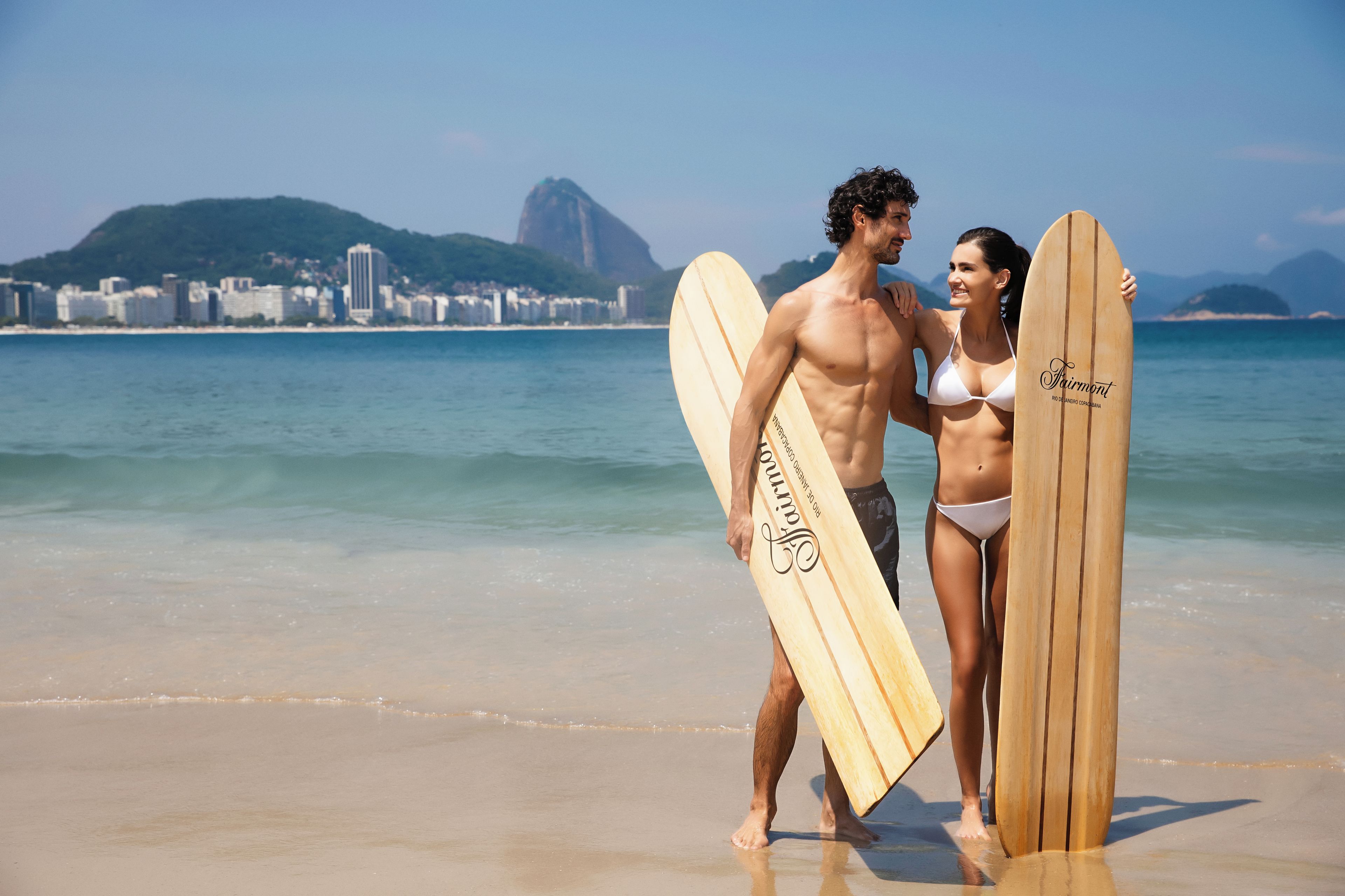 A beach scene with a couple holding surfboards, a clear sky, and a cityscape in the background.