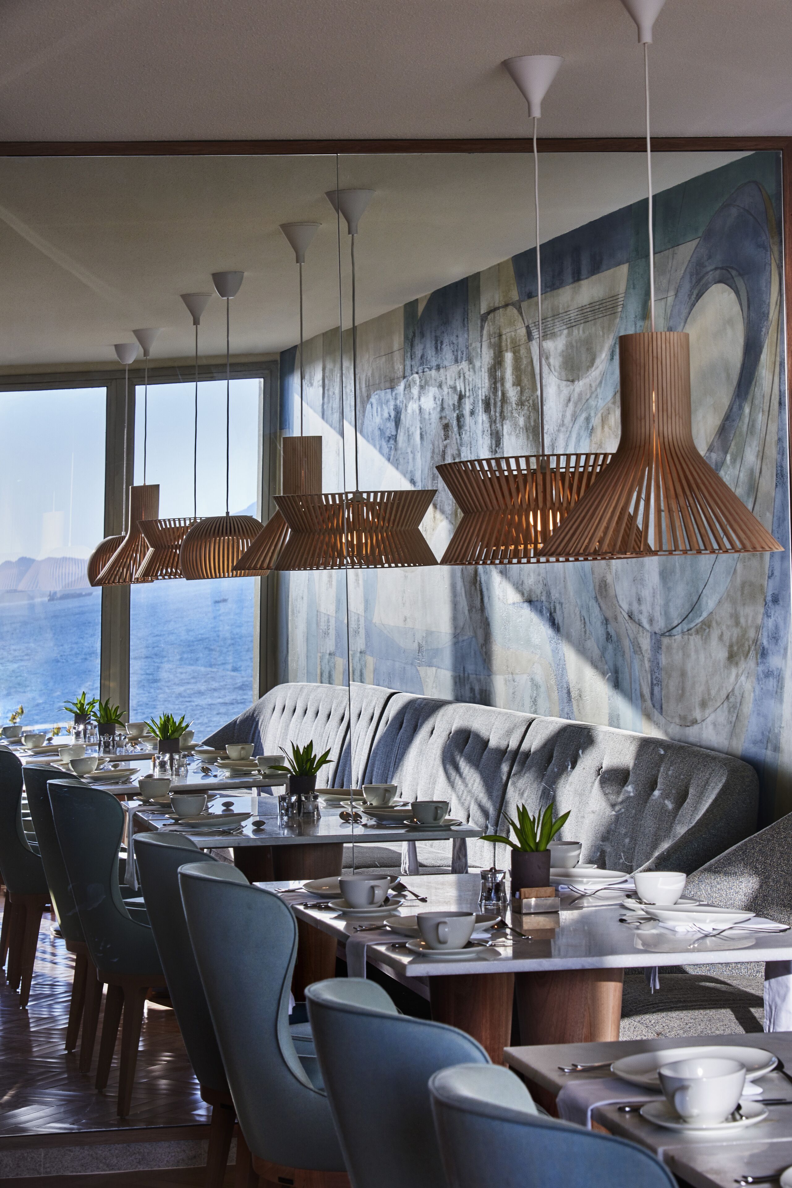 Un comedor con vista al mar, que cuenta con una mesa grande dispuesta para comer, iluminación colgante moderna y un cuadro abstracto de gran tamaño en la pared.