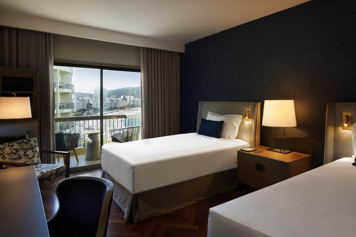 Ein Hotelzimmer mit zwei Betten, einem Schreibtisch, einem Sessel, einem Fernseher und einem großen Fenster mit Blick auf die Stadt.