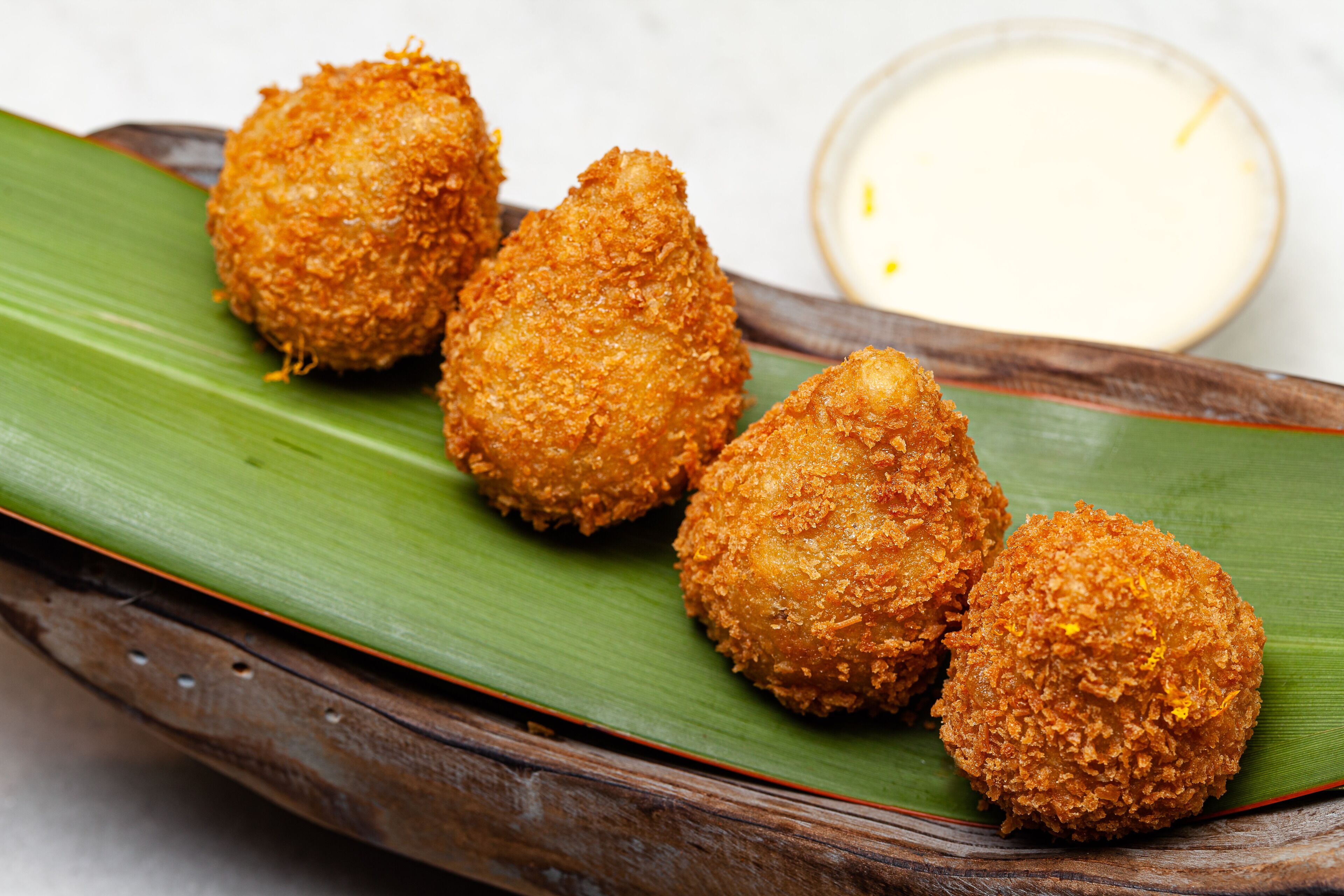 Four breaded and fried balls on a green leaf, with a small bowl of dipping sauce beside them.