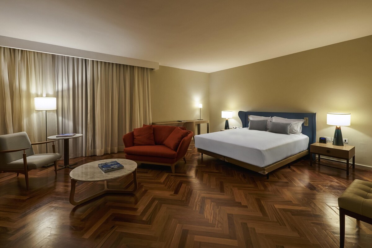 A hotel room with a bed, two chairs, a small table, a desk, and a lamp.