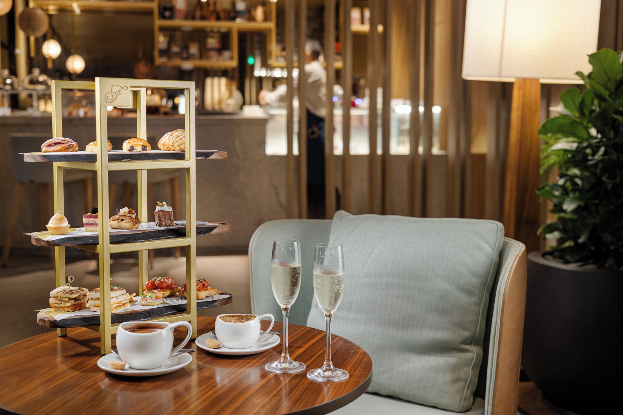 Uma área de refeições com uma mesa de sobremesas em níveis, uma mesa redonda com uma xícara de café e duas taças de champanhe, e uma poltrona confortável.