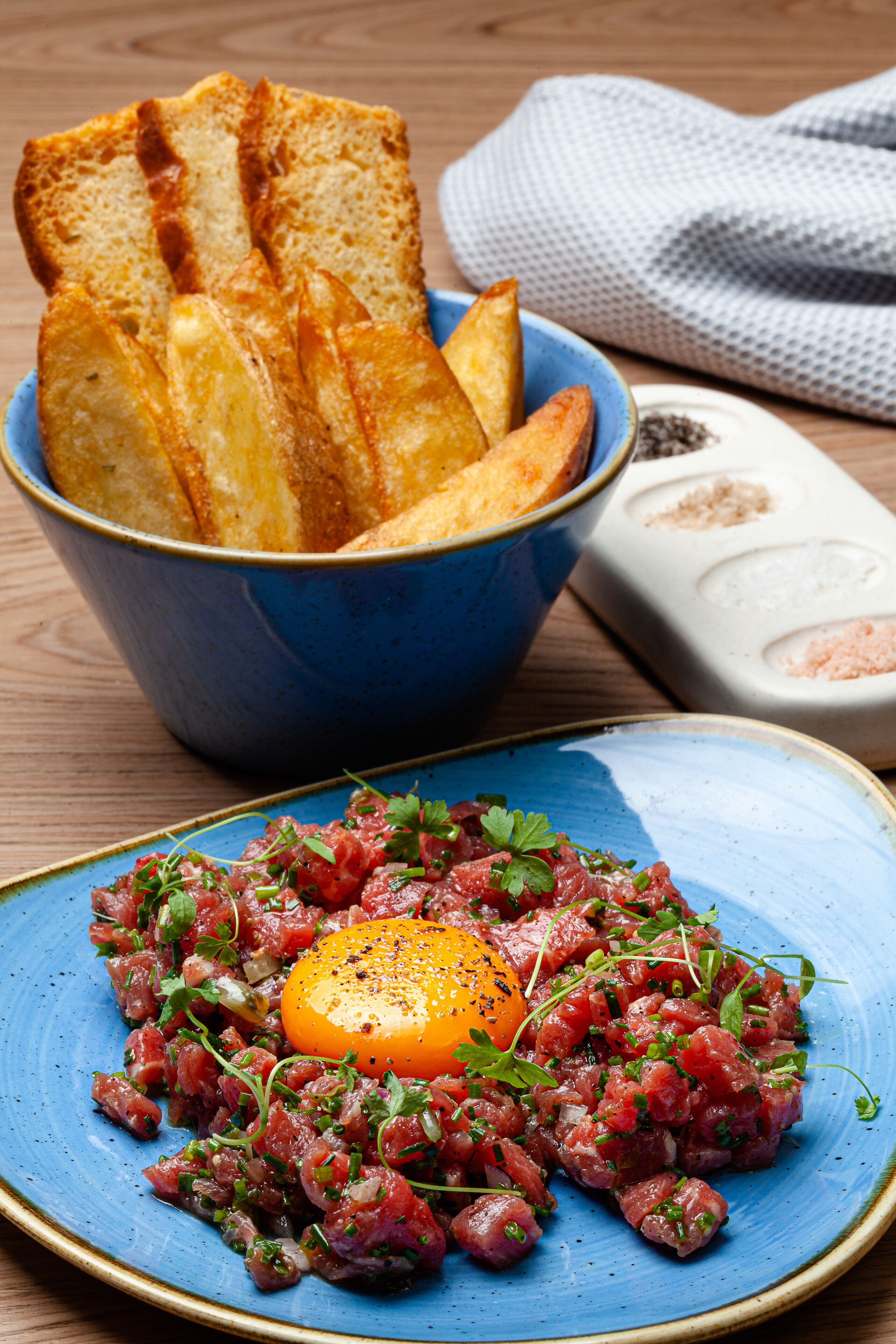 A plate of steak tartare with a raw egg yolk, garnished with herbs, and a bowl of toasted bread in the background.