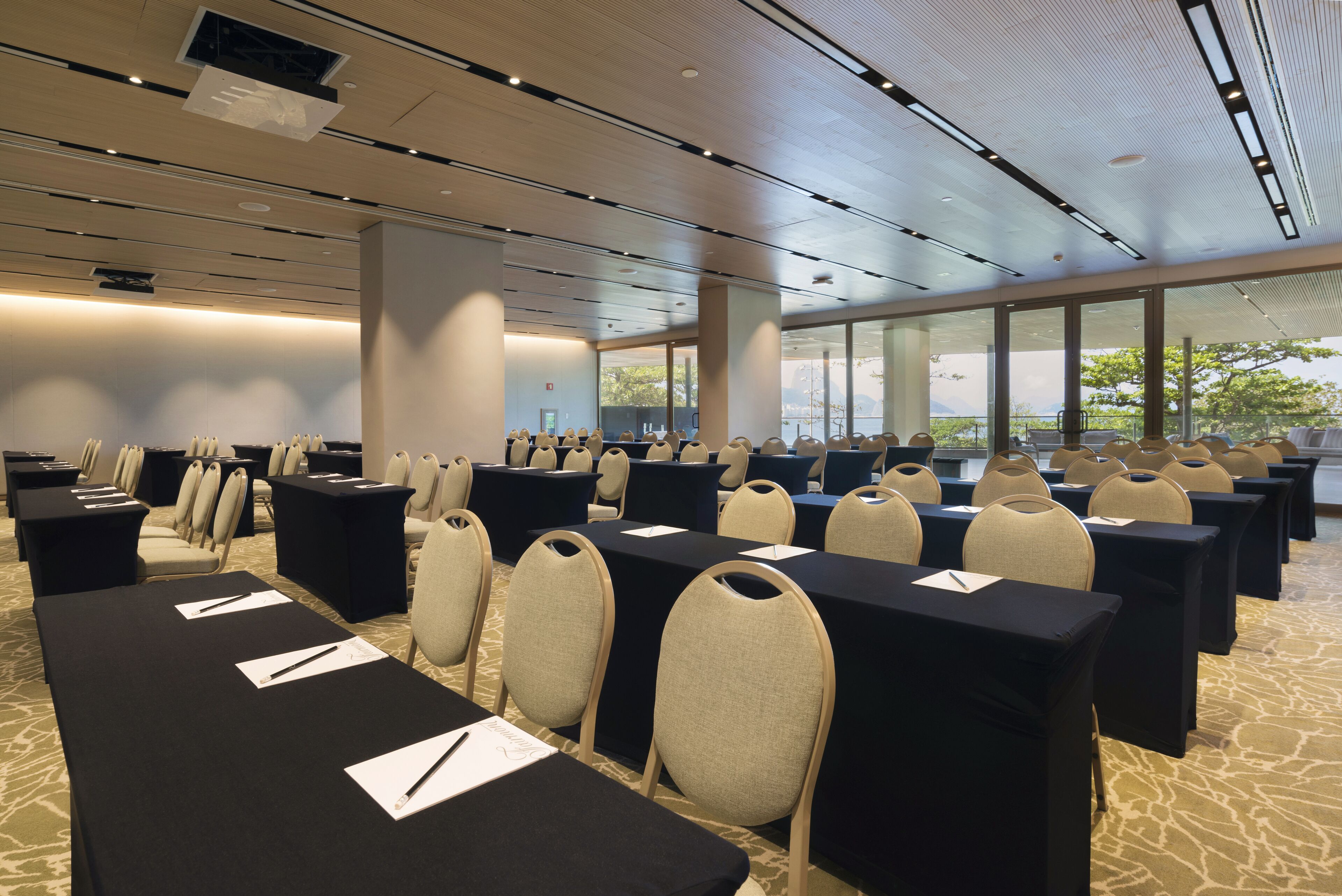 Une salle de conférence avec des rangées de tables et de chaises, de grandes fenêtres et un plafond doté d’un éclairage encastré.
