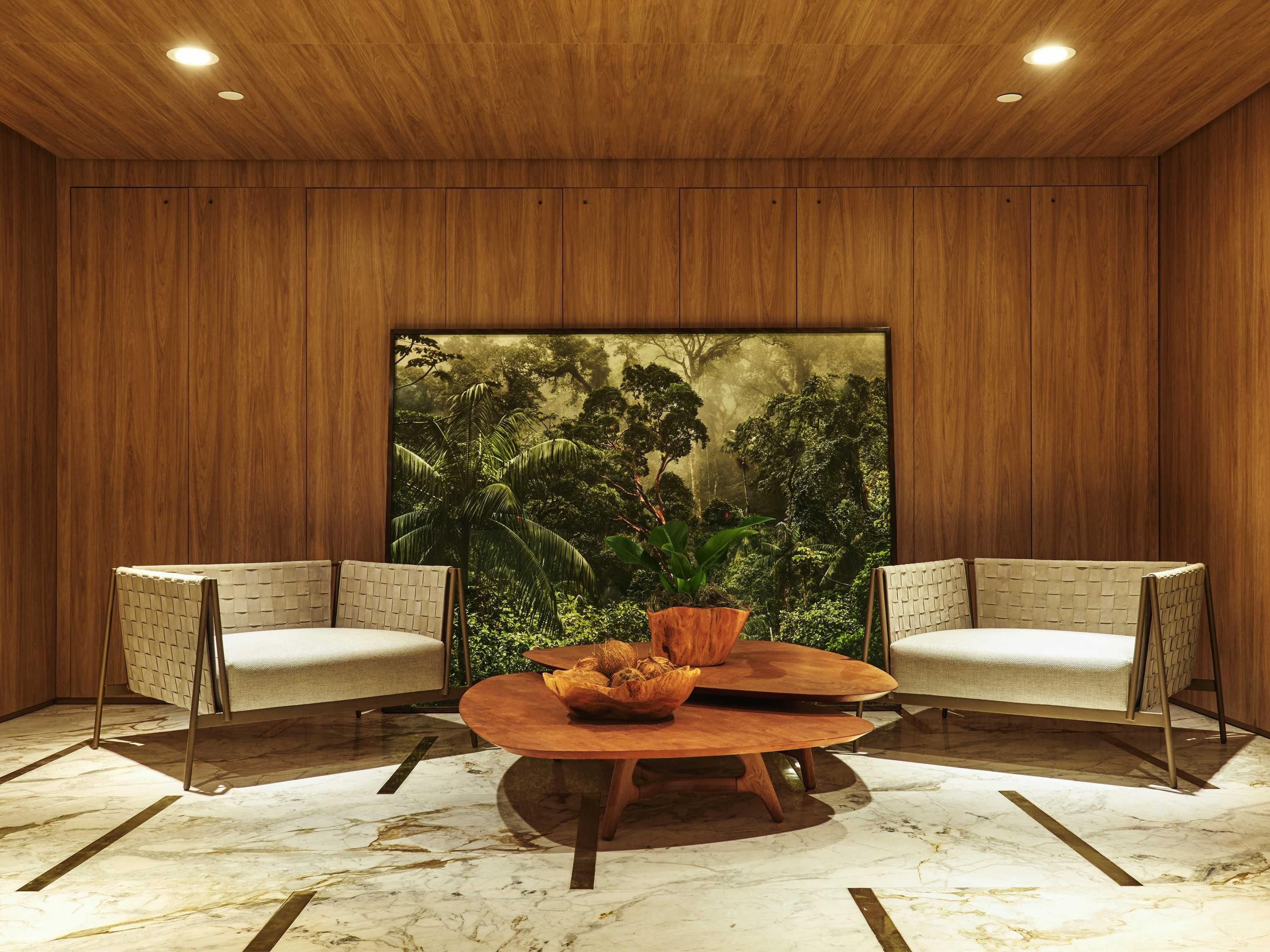 A room with wooden panel walls, a large forest-themed artwork, two cushioned chairs, a round wooden table with a bowl and plant, and a marble floor.