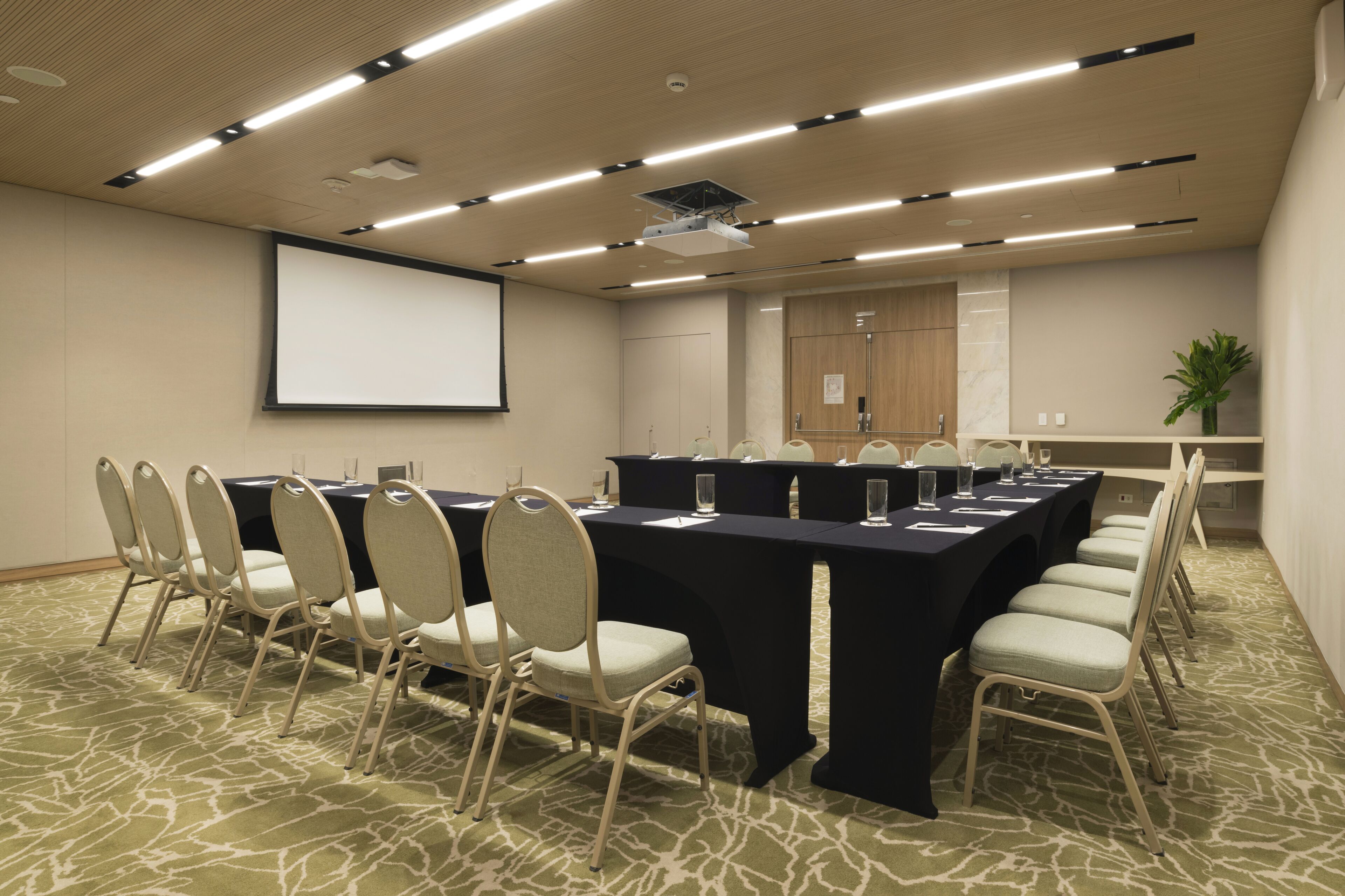 Un salón de conferencias con una larga mesa, sillas, una pantalla de proyector y suelo alfombrado.