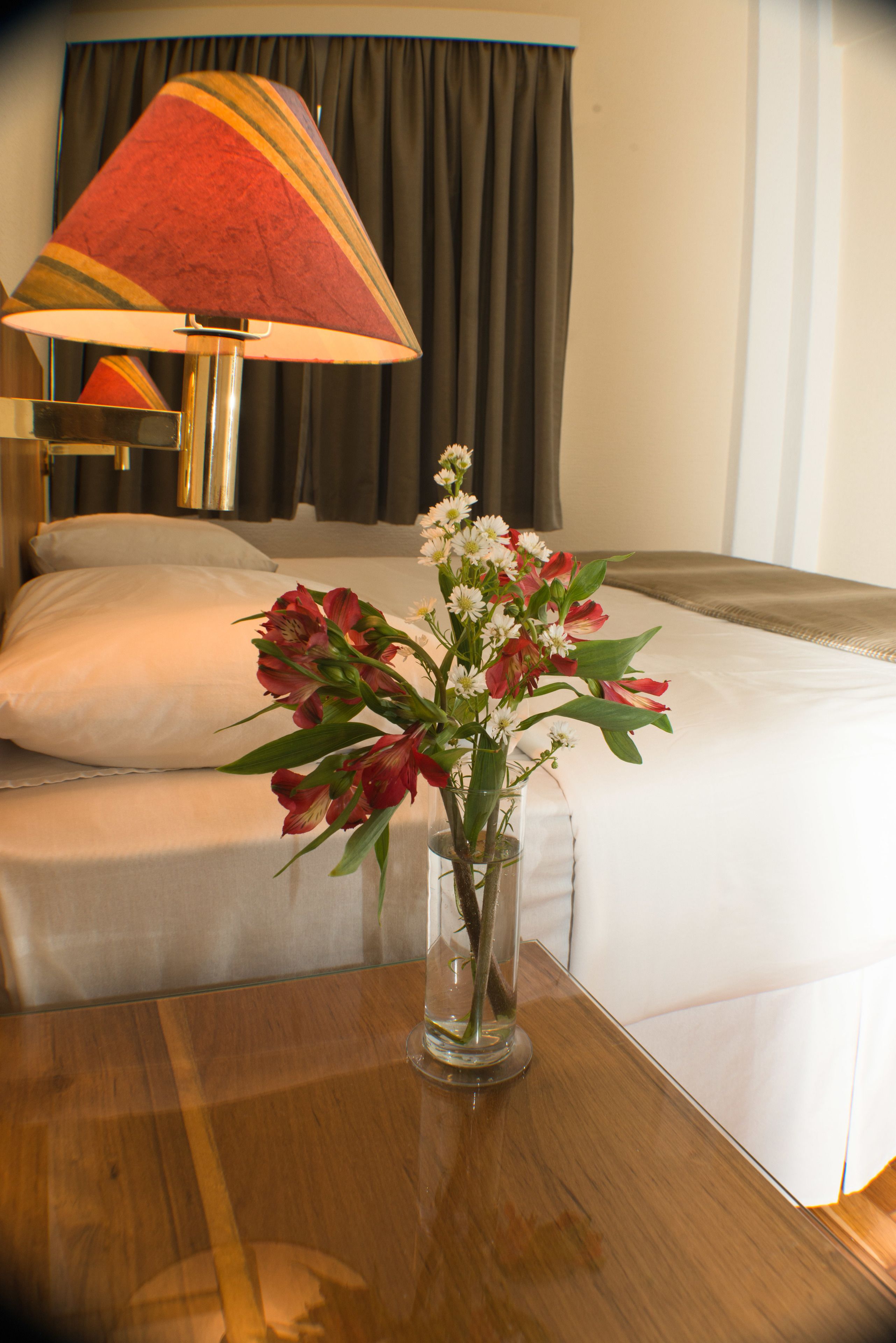 A hotel room with a bed, a nightstand with a lamp, and a vase with flowers on the table.