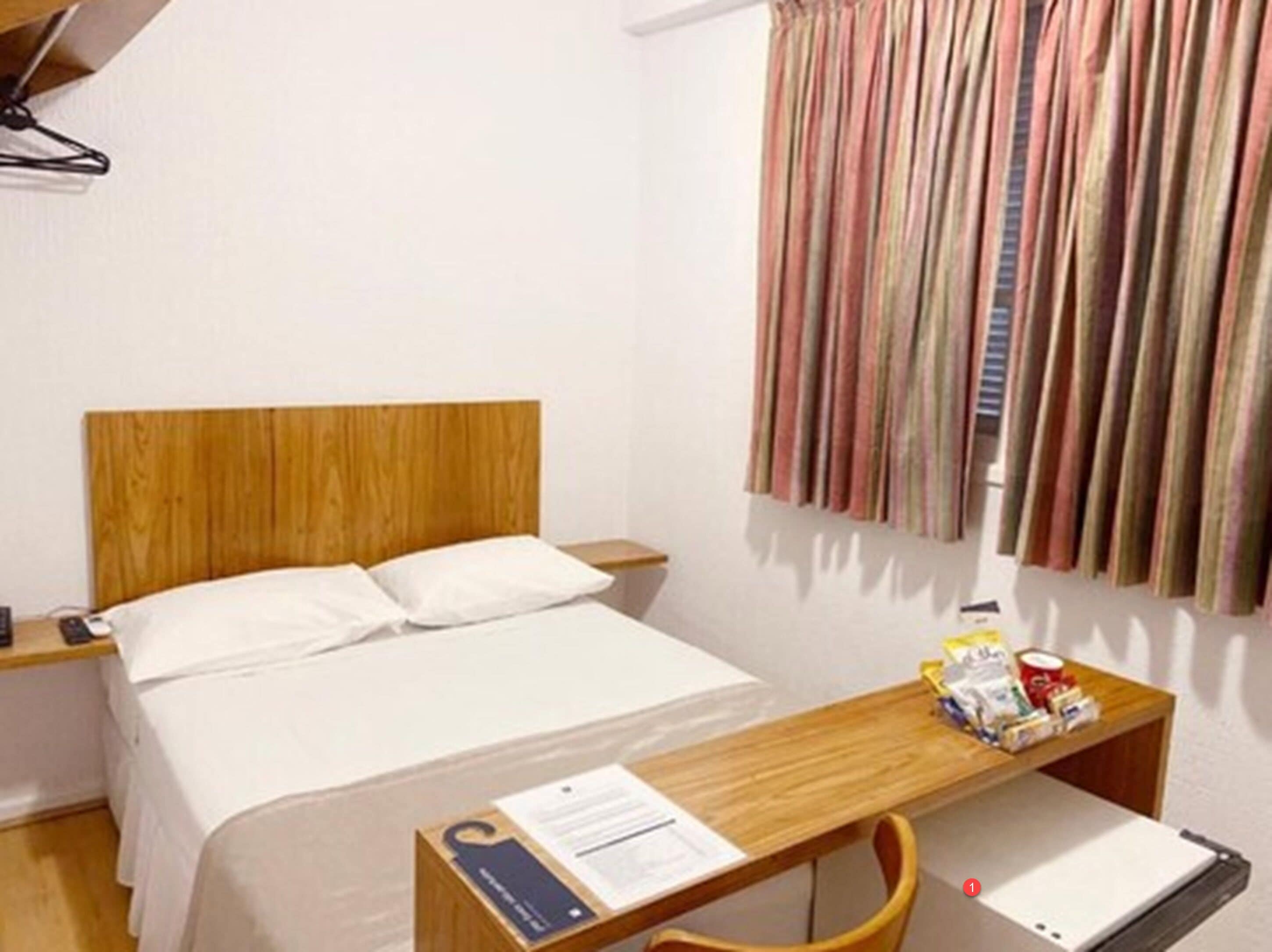 Une chambre d’hôtel avec un lit doté d’une tête de lit en bois, une literie blanche, une table de chevet en bois avec une lampe, un bureau avec une chaise et un rideau à rayures.