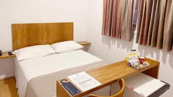A hotel room with a wooden headboard bed, white bedding, a wooden nightstand with a lamp, a desk with a chair, and a striped curtain.