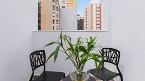 Un pequeño balcón con dos sillas negras y una mesa, con vista a edificios de la ciudad.